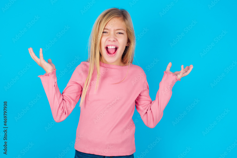 Crazy outraged caucasian little kid girl wearing long sleeve shirt over blue background screams loudly and gestures angrily yells furiously. Negative human emotions feelings concept Crazy outraged caucasian little kid girl wearing long sleeve shirt over blue background screams loudly and gestures angrily yells furiously. Negative human emotions feelings concept