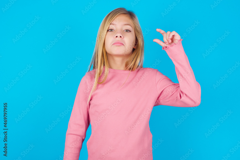Fototapeta premium caucasian little kid girl wearing long sleeve shirt over blue background purses lip and gestures with hand, shows something very little.