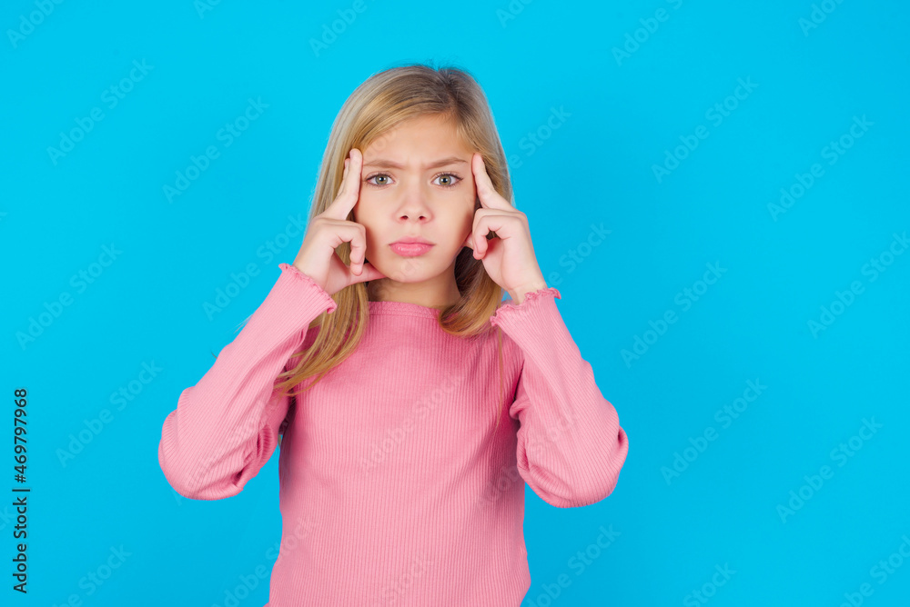 Serious concentrated caucasian little kid girl wearing long sleeve shirt over blue wall keeps fingers on temples, tries to ease tension, gather with thoughts and remember important information