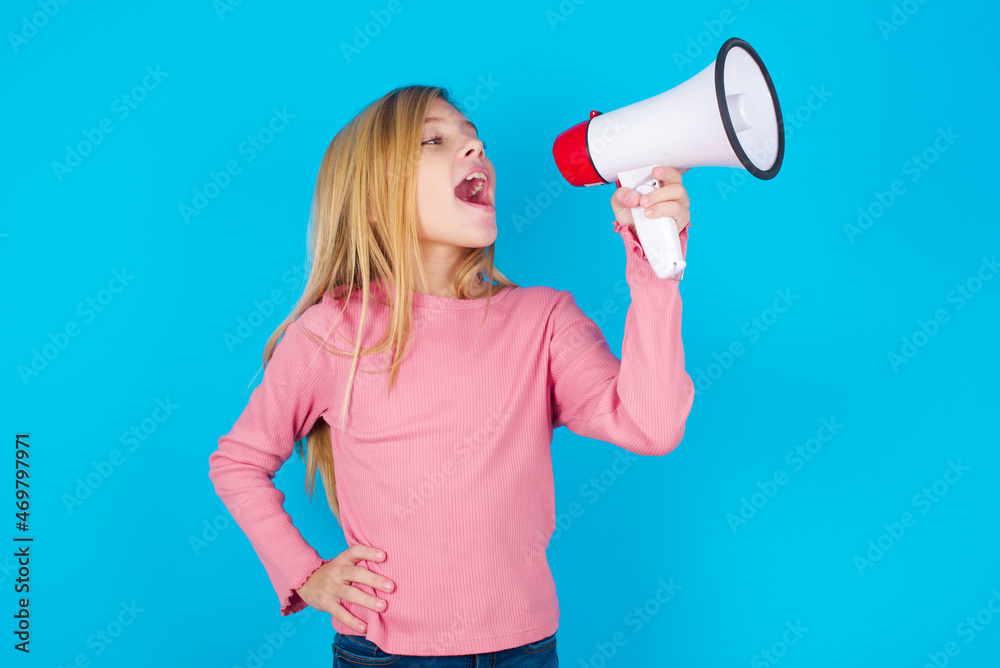Funny caucasian little kid girl wearing long sleeve shirt over blue background People sincere emotions lifestyle concept. Mock up copy space. Screaming in megaphone.