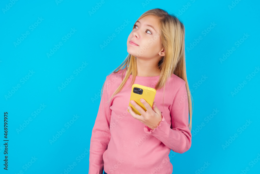 caucasian little kid girl wearing long sleeve shirt over blue background holds mobile phone uses high speed internet and social networks has online communication. Modern technologies concept caucasian little kid girl wearing long sleeve shirt over blue background holds mobile phone uses high speed internet and social networks has online communication. Modern technologies concept