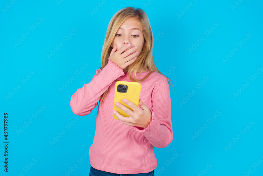 caucasian little kid girl wearing long sleeve shirt over blue background being deeply surprised, stares at smartphone display, reads shocking news on website, Omg, its horrible! caucasian little kid girl wearing long sleeve shirt over blue background being deeply surprised, stares at smartphone display, reads shocking news on website, Omg, its horrible!