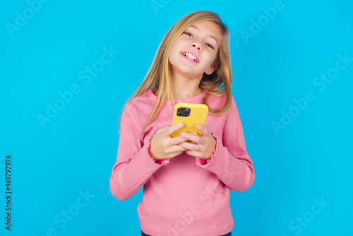 caucasian little kid girl wearing long sleeve shirt over blue background Mock up copy space. Using mobile phone, typing sms message