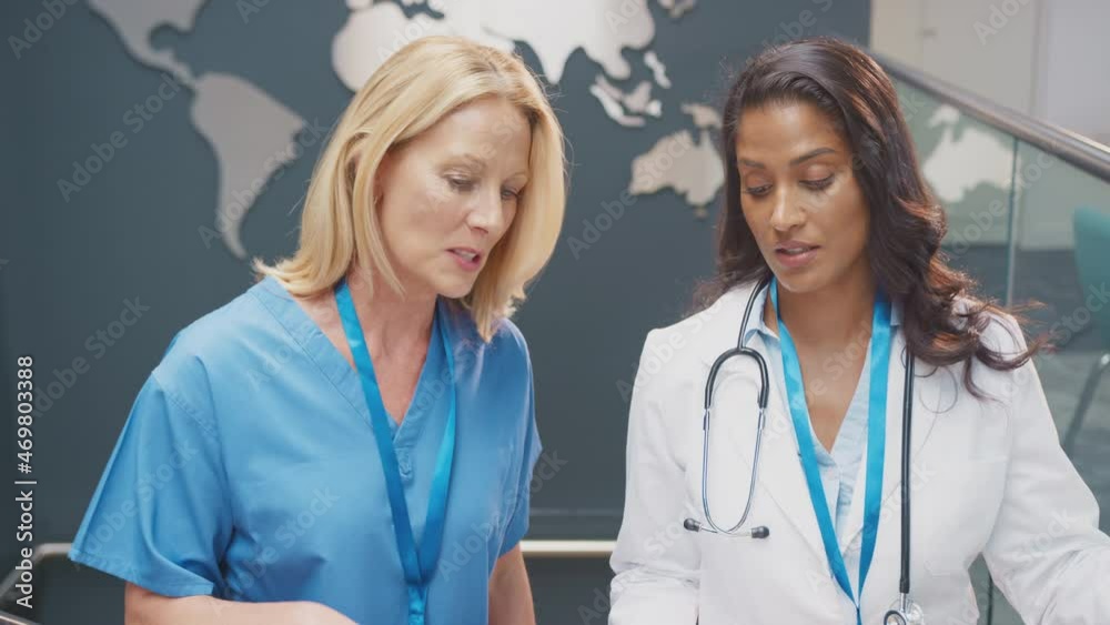 Two female doctors in white coat and scrubs discussing patient notes during Informal meeting in hospital with world map in background - shot in slow motion Two female doctors in white coat and scrubs discussing patient notes during Informal meeting in hospital with world map in background - shot in slow motion