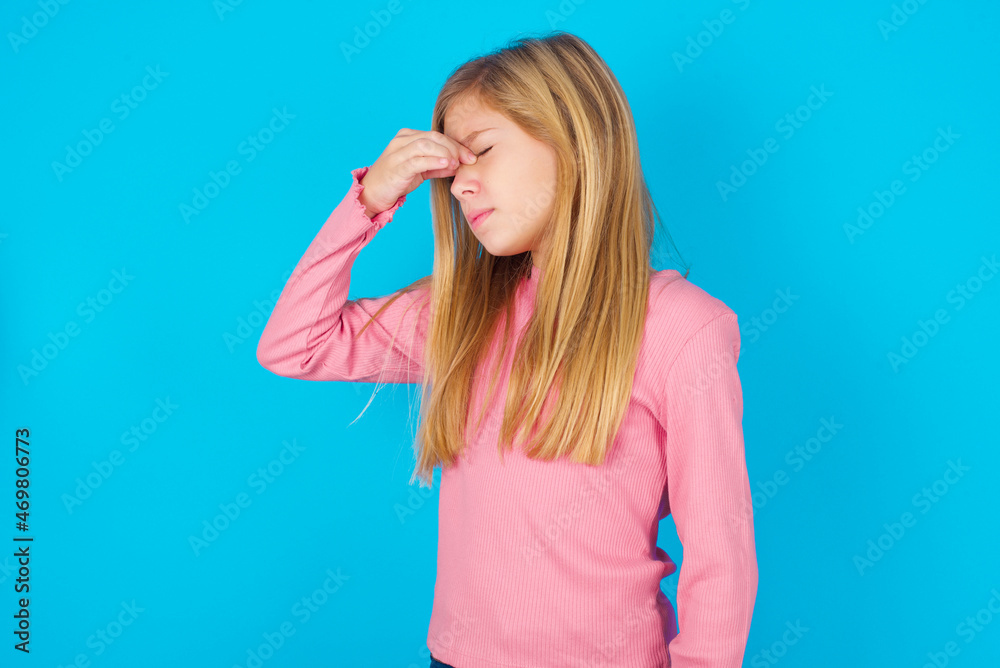 Sad caucasian little kid girl wearing long sleeve shirt over blue background suffering from headache holding hand on her face