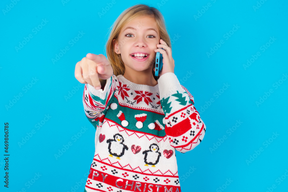 Positive caucasian little kid girl wearing knitted sweater christmas over blue background indicates directly at camera has telephone conversation smiles broadly enjoys talking long hours. You join me Positive caucasian little kid girl wearing knitted sweater christmas over blue background indicates directly at camera has telephone conversation smiles broadly enjoys talking long hours. You join me