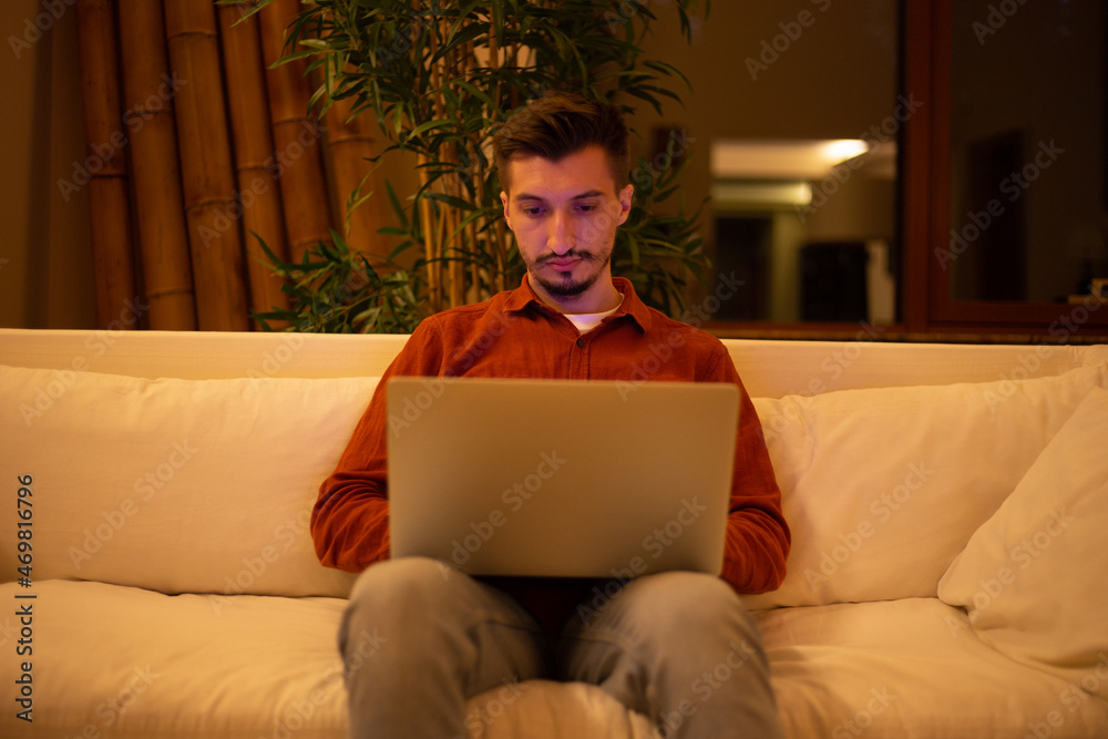 A young man with a beard in a red shirt works with a laptop and sits on the couch in the evening in the house. A young man with a beard in a red shirt works with a laptop and sits on the couch in the evening in the house.
