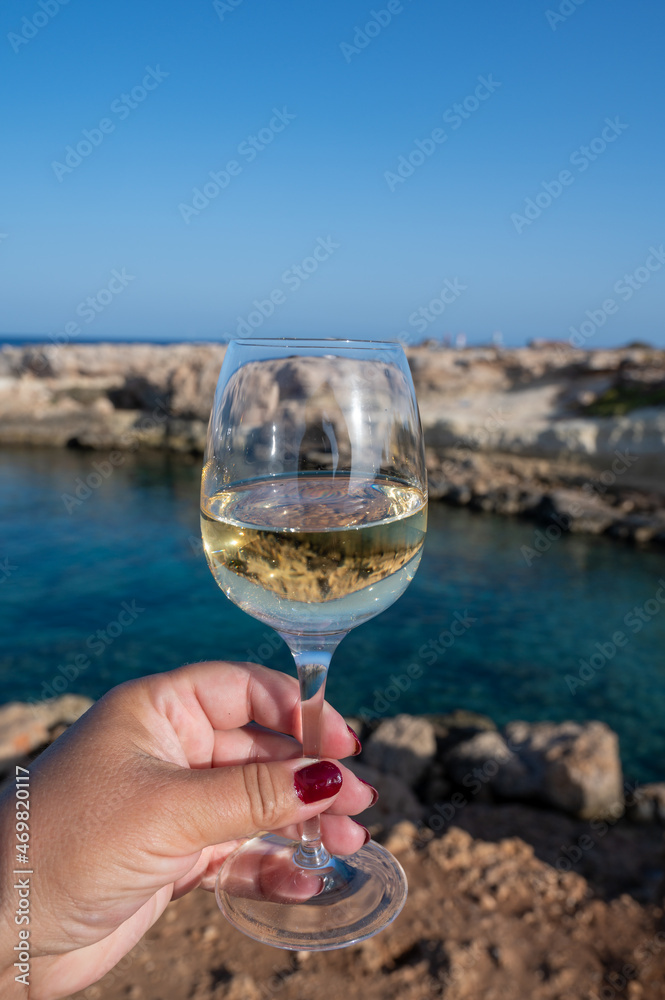 Woman's hand holding glass of white dry white wine with view on rocks and blue sea bay water near Protaras touristic town on Cyprus