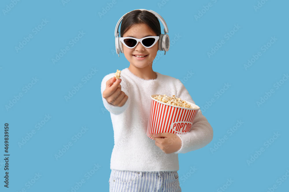 Fototapeta premium Happy cute girl in headphones and sunglasses holding bucket of popcorn on blue background