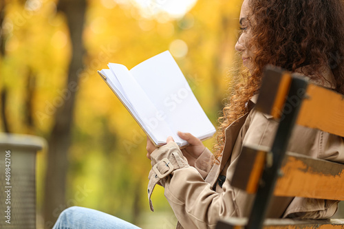 Young African-American woman reading book with blank pages on bench in autumn park