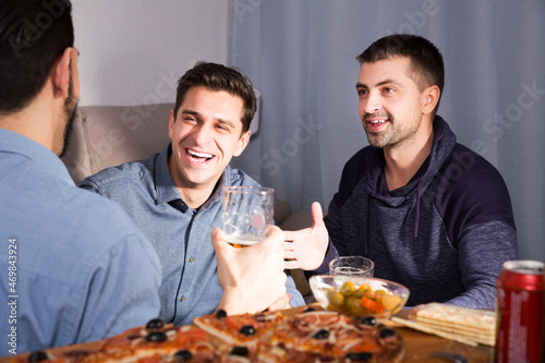 Three adult glad men talking and laughing while enjoying beer and pizza at home