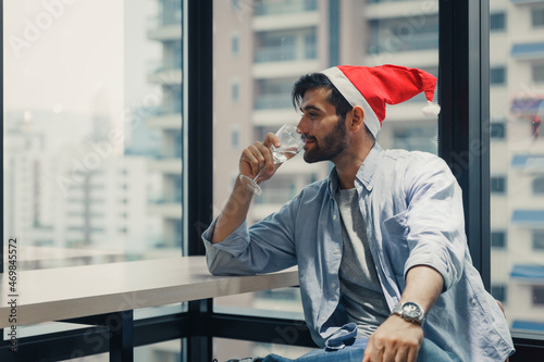 Attractive Caucasian man drinking champagne from the glass sitting against big building view. Young handsome men wearing red hat celebrating x'mas and happy new year alone