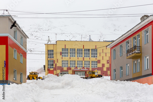 Snow-covered street in a northern town in the Arctic. Bulldozers remove snow. Clearing snow from streets after a blizzard. Large snowdrifts near buildings. Provideniya, Chukotka, Far North of Russia. © Andrei Stepanov