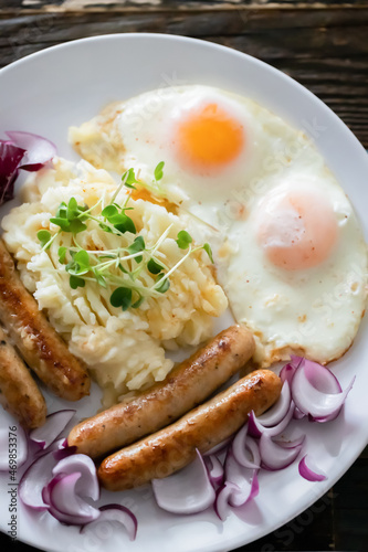 Pork sausages with onions, eggs, mashed potatoes and microgreens. Hearty breakfast. Dark wooden background, white ceramic plate. Top view.
