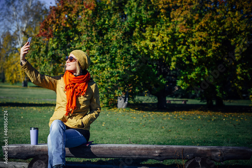 Cheerful woman in sunglasses, sitting on wooden bench in autumn park, taking selfie on smartphone. Blurred green background, copy space.