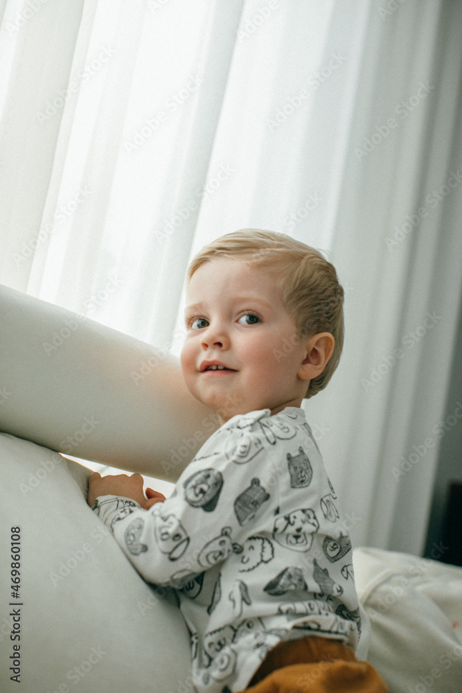 An incredibly handsome boy with blond hair is playing on a soft white sofa. Vertical photography