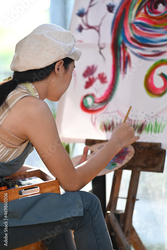 Side view young woman artist sitting in front of the canvas and painting with water color.
