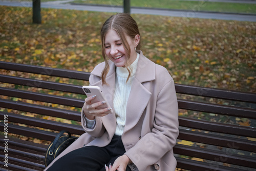 A young girl in a beautiful stylish coat sits on a bench in the city with her hair gathered, holds a smart phone in her hands and laughs