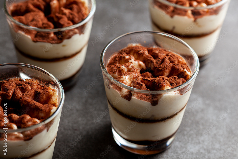 Delicious traditional tiramisu desert served in small glass cups on stone countertop. Low light image shows texture and layers from angle with shallow depth of field. Delicious traditional tiramisu desert served in small glass cups on stone countertop. Low light image shows texture and layers from angle with shallow depth of field.