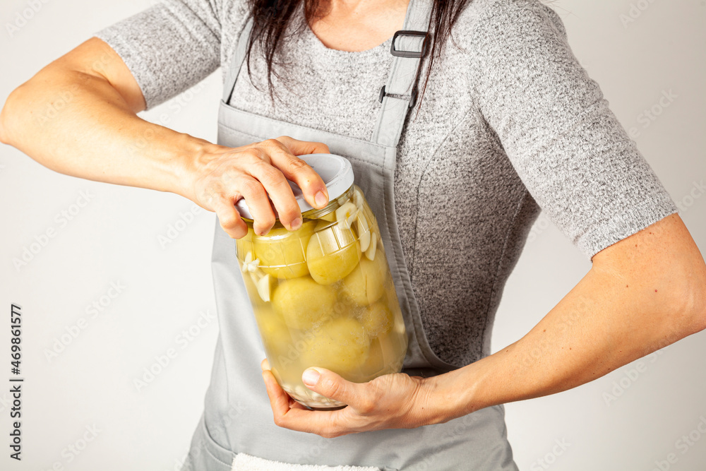 A caucasian woman chef wearing apron is trying to open a stubborn jar lid. She uses force to unscrew the lid in kitchen. Concept image for pickling tomatoes and gherkins and difficulty in opening lids - obrazy, fototapety, plakaty 