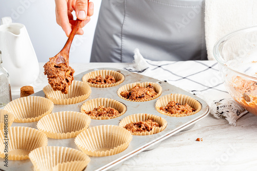 making of delicious fruit and nut cupcake on white marble countertop background. Muffin tin with liner, ingredients, and utensils are seen. A woman is adding mixture into the holes
