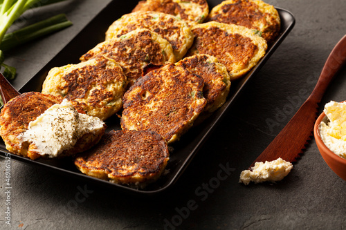 Turkish style zucchini pancakes or fritters known as mucver, served on black plate with fresh greens and sour cream. Close up image on dark stone background.