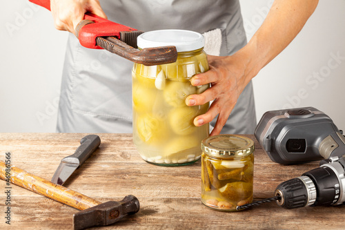A funny quirky concept image showing a caucasian woman trying to open a jar of pickles. She is trying with tools like drill, knife, wrench and hammer