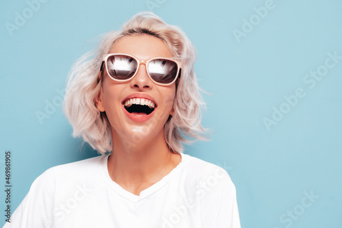 Portrait of young beautiful smiling female in trendy summer white clothes.Sexy carefree blond woman posing near blue wall in studio. Positive model having fun indoors. Cheerful and happy in sunglasses © halayalex