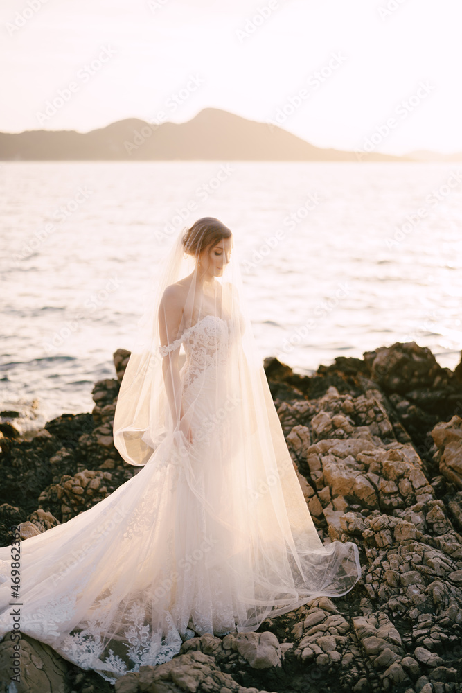 Bride in a white dress and veils stands on the rocks by the sea