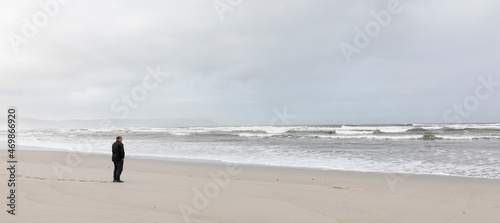 A man walking across sand to the water's edge on a beach, overcast day and surf waves breaking on shore. 