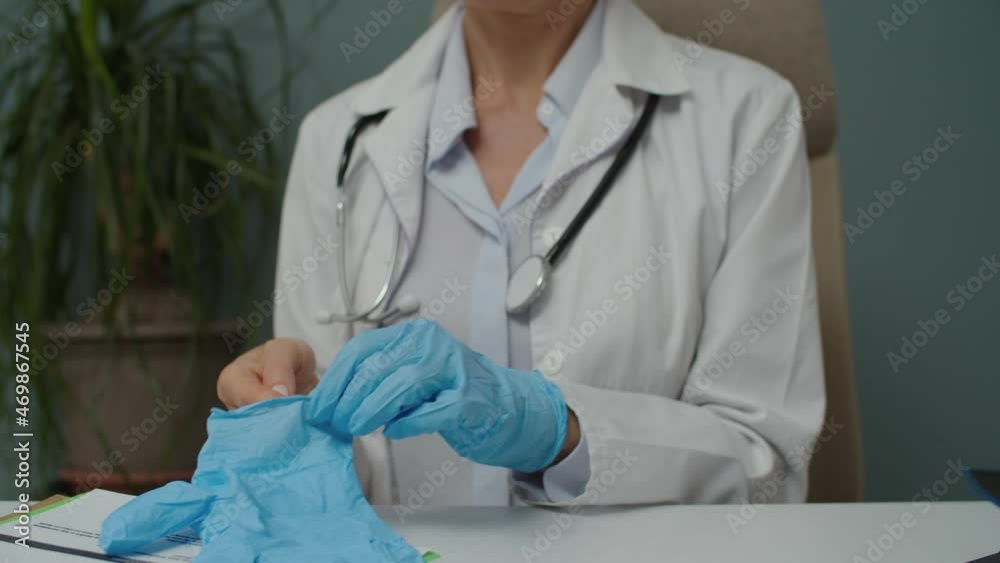 Midsection of unrecognizable female doctor in white lab coat putting on blue medical protective gloves indoors. MD woman preparing for treatment or procedure, applying sterile latex gloves