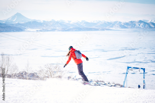 Snowboarder girl in red jacket riding down the hill in front of snowy mountains and blue sky