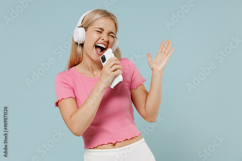 Young happy caucasian blonde woman 20s in casual pink t-shirt headphones listen to music sing song in microphone record voice on mobile phone dictaphone isolated on plain pastel light blue background.