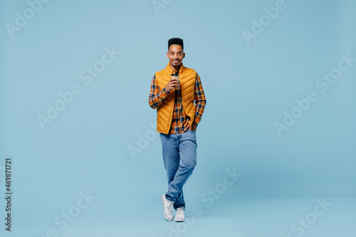 Full size body length smiling young black man 20s wears yellow waistcoat shirt hold takeaway delivery craft paper brown cup coffee to go isolated on plain pastel light blue background studio portrait.