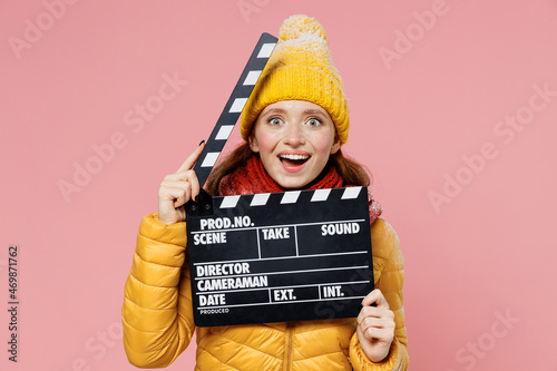 Overjoyed excited young woman 20s years old wear yellow jacket hat mittens looking camera holding classic black film making clapperboard isolated on plain pastel light pink background studio portrait © ViDi Studio