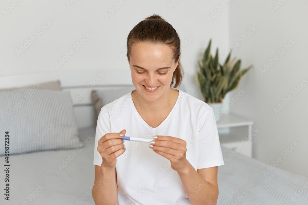 Extremely happy attractive woman wearing casual attire sitting on sofa at home and looking at pregnancy test with positive result in her hands, expressing positive emotions. Extremely happy attractive woman wearing casual attire sitting on sofa at home and looking at pregnancy test with positive result in her hands, expressing positive emotions.