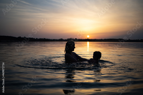 Mom plays with a naked baby in oversleeves in the lake against the background of a summer sunset