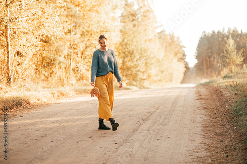 Photo of young woman stopped her car while traveling, went out on village road and strolled along it, enjoying autumn beauty of the trees, the rays of the setting sun smiling happily.