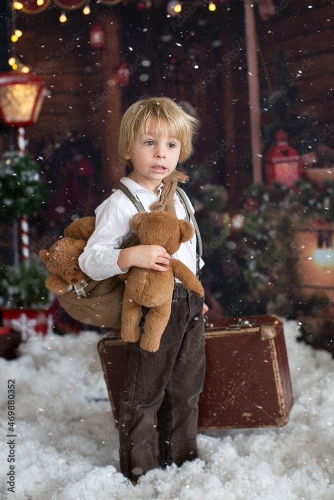 Cute fashion toddler boy, playing in the snow with teddy bear in front of a wooden cabin log