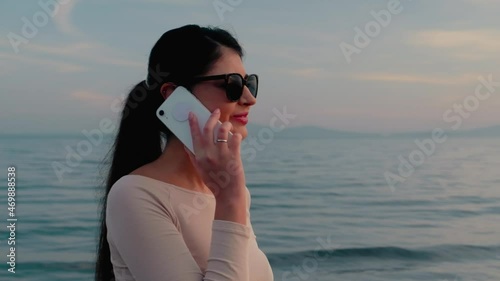 Happy young brunette girl in glasses on the background of the sea communicates using a mobile phone