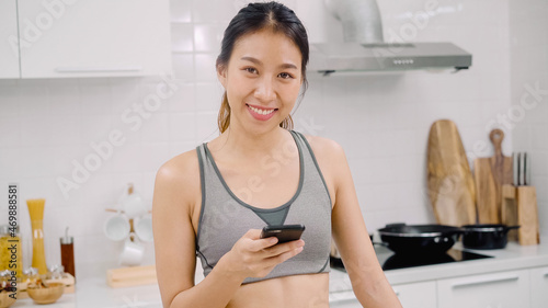 Young Asian blogger woman using smartphone for talking, chatting and checking social media in the kitchen, female in sport clothing cooking at home. Lifestyle women relax at home concept.