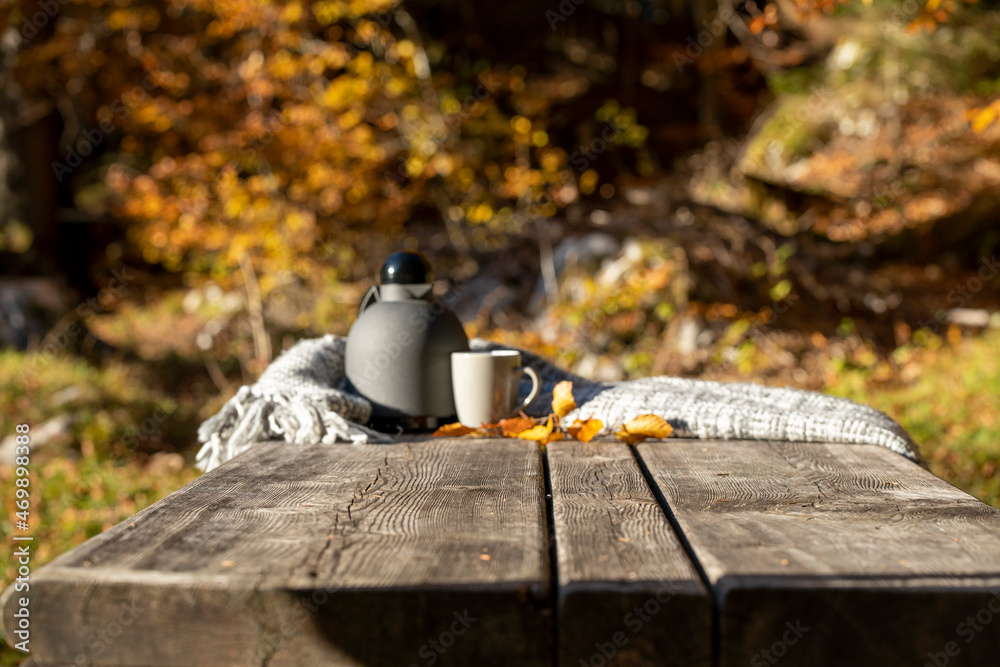 there is a thermos and a mug of hot tea with a gray blanket and yellow leaves on a wooden table in autumn in nature