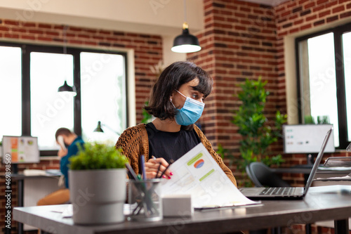 Executive manager with medical protection face mask to prevent infection with covid19 analyzing business graphs typing management strategy on laptop computer. Businesswoman working at project ideas