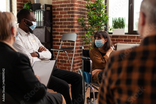 People and woman sitting in wheelchair having conversation at aa meeting. Multi ethnic patients attending conversation with psychiatrist at support group therapy during covid 19 pandemic.