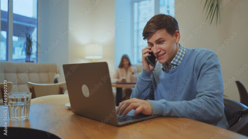 Successful businessman wearing sweater sitting in cafe, talking on phone and using laptop, woman on blurred background. Tracking shot of young man working remotely. Concept of freelance Successful businessman wearing sweater sitting in cafe, talking on phone and using laptop, woman on blurred background. Tracking shot of young man working remotely. Concept of freelance