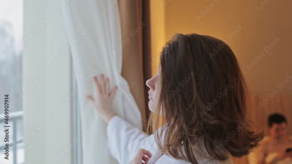 A young woman, opening the curtains, looks out the window on a winter morning. A young man in the background in bed using the phone. Weekend in a country hotel, winter vacation concept A young woman, opening the curtains, looks out the window on a winter morning. A young man in the background in bed using the phone. Weekend in a country hotel, winter vacation concept