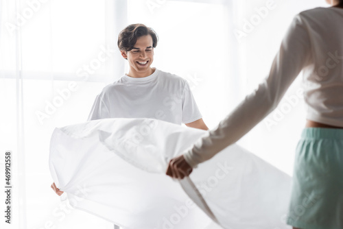 happy man arranging bed sheet with african american girlfriend