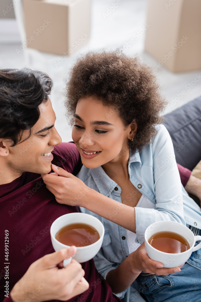 pleased multiethnic couple holding cups of tea while resting on couch pleased multiethnic couple holding cups of tea while resting on couch