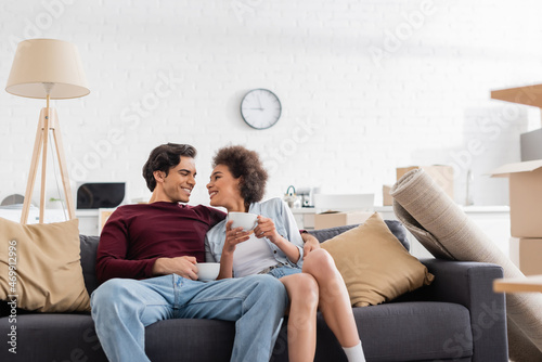 happy multiethnic couple holding cups of tea while resting on couch during relocation