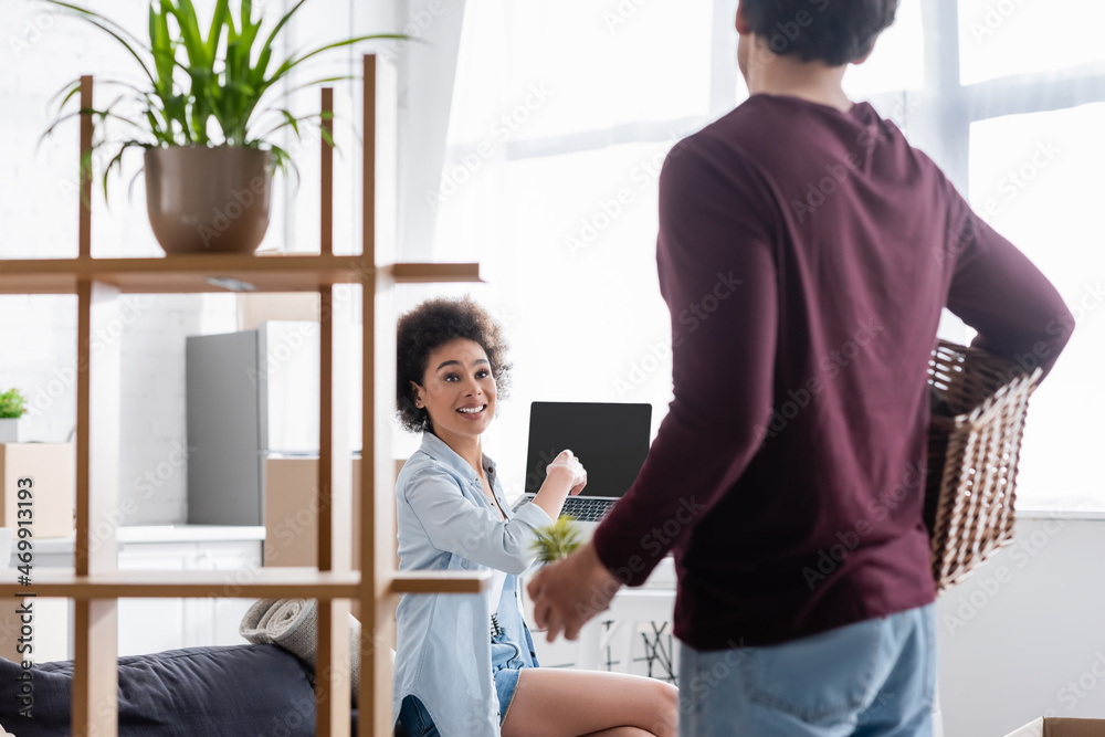 happy african american woman pointing at laptop while looking at blurred man holding plant and basket happy african american woman pointing at laptop while looking at blurred man holding plant and basket